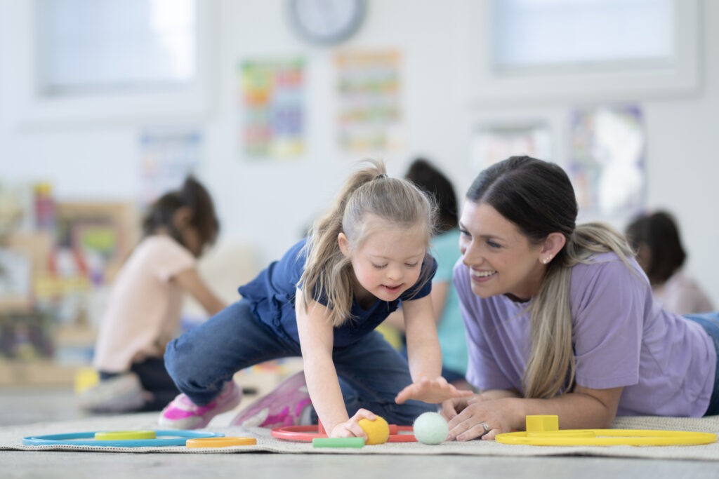 A sweet little girl with Downs Syndrome works with her teacher on the floor of the classroom as they short toys by colour.  The two are both dressed comfortably and focused on the task as they work together.